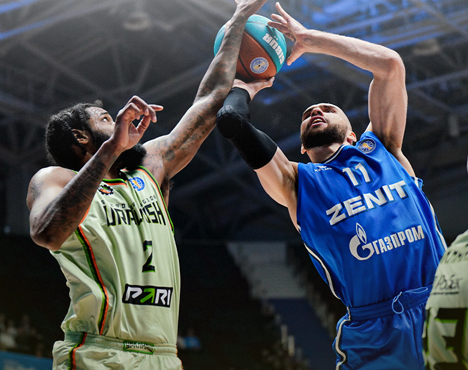 Два баскетболиста борются за мяч под кольцом. Игрок слева в зелёной форме команды Уникс, игрок справа в синей форме команды Зенит. Атмосфера игры напряжённая, на заднем плане видны трибуны
