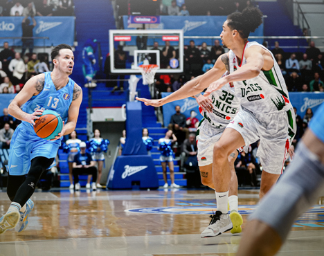 На изображении баскетболисты на площадке в разгар игры. Игрок в голубой форме с мячом атакует, а соперник в белой форме с зеленым узором пытается защититься. На заднем плане видны болельщики и часть трибун