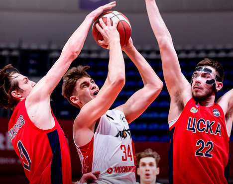 Баскетбольный матч: игрок в белой форме пытается забросить мяч в кольцо, находясь под прессингом двух соперников в красной форме. Игроки демонстрируют напряжённую борьбу за мяч