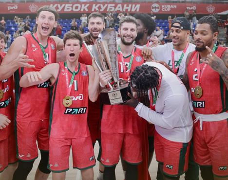Группа баскетболистов в красной форме празднует победу, держа трофей. Некоторые игроки в медалях выглядят радостными и удивленными. На заднем плане видно трибуны с людьми