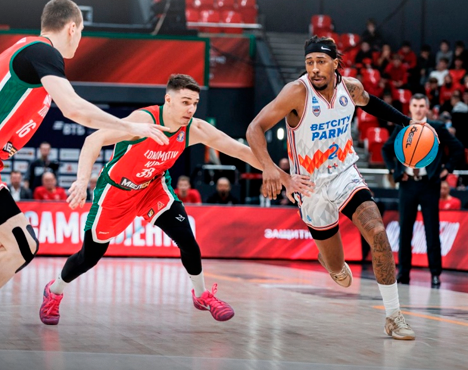 На изображении запечатлен момент баскетбольного матча: игрок в белой форме с мячом прорывается между двумя соперниками в красно-зеленой форме на яркой спортивной арене